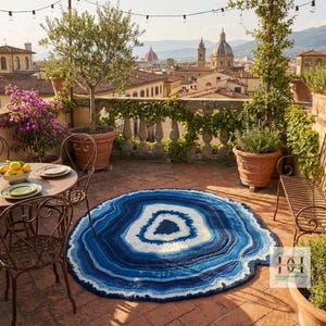 May include: A circular rug with an agate design in shades of blue and white is placed on a brick patio. The rug's pattern consists of concentric rings of varying blue hues, with a white central area. The patio overlooks a cityscape.