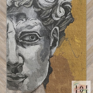 Op de afbeelding: Een rechthoekig tapijt met een gedetailleerde, grijstinten weergave van het gezicht van een klassiek beeldhouwwerk, tegen een goudkleurige achtergrond. Het ontwerp omvat het oog, de neus en de lippen van het beeldhouwwerk. Het tapijt is een decoratief item.