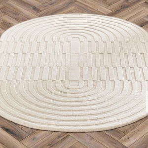Op de afbeelding: Crème-kleurig, ovaal tapijt met een geometrisch patroon. Het tapijt heeft concentrische halve cirkels en rechthoekige vormen, waardoor een textuur ontstaat. Het ligt op een houten visgraatvloer.