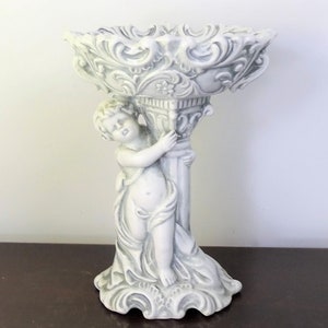 May include: A white and gray cherub statue holding a decorative bowl. The statue has intricate details and a classical design. The bowl has a detailed, ornate rim. The statue is on a dark brown surface.
