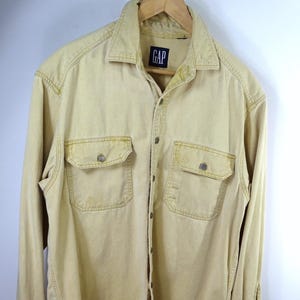 May include: A light yellow button-down shirt with two chest pockets and the GAP logo on the label. The long-sleeved shirt is made of a sturdy fabric and has buttoned cuffs. The shirt is hanging on a wooden hanger.
