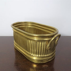 May include: A brass planter with a rectangular oval shape and vertical ribbed detailing. The planter has a curved handle on each end and a smooth rim. The brass has a shiny, golden finish. The planter sits on a dark brown surface.