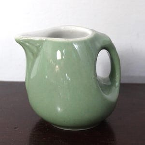 May include: A small, sage green ceramic creamer pitcher with a rounded body, a short pouring spout, and a curved handle. The interior of the pitcher is white, and the exterior features a smooth, glossy finish with a consistent light green color.