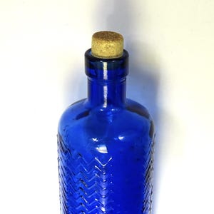 May include: A cobalt blue glass bottle with a textured chevron pattern and a cork stopper. The bottle has a rounded shape and a narrow neck. The cork is a light brown color. The bottle is likely used for storing liquids.