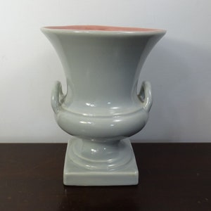 May include: A light gray ceramic urn with a square base and two curved handles. The interior rim is a soft pink. The urn has a classic, symmetrical design, suitable for decorative purposes or displaying flowers. The urn is a light gray color.
