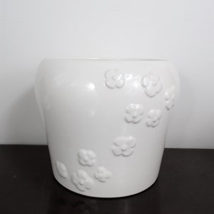 May include: A white ceramic flower pot with a glossy finish. The pot features a series of raised, three-dimensional flower designs scattered across its surface. The pot is cylindrical with a slightly curved shape.