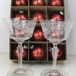 Op de afbeelding: Twee heldere, geëtste champagnecoupeglazen met bloemmotieven. De glazen staan voor een kartonnen doos gevuld met rode kerstballen. De ballen zijn rond en glanzend.