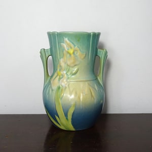 May include: A ceramic vase featuring a gradient color scheme transitioning from deep blue at the base to light green and yellow at the top. The vessel has two side handles and a raised relief design of iris flowers with yellow centers.