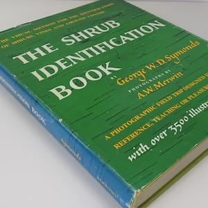 Puede incluir: Un libro antiguo titulado "The Shrub Identification Book" de George W.D. Symonds. La portada es verde con texto blanco y amarillo, incluyendo "over 3500 illustrations". El lomo del libro es azul con el nombre del autor.