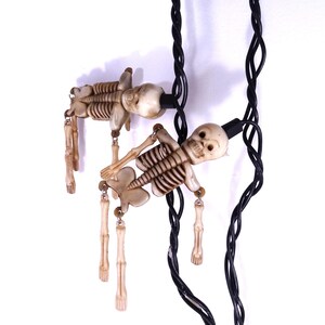 May include: A string of decorative lights featuring two off-white, molded plastic skeleton figures. Each skeleton has articulated limbs attached by small metal rings to a black electrical cord, with a black plastic light socket housing.