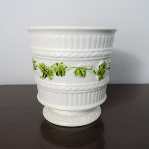 Puede incluir: Jardinera de cerámica blanca con base de pedestal y diseño texturizado. La jardinera presenta estrías verticales, bandas tipo cuerda y un motivo de vid y hojas verdes en el centro. Una pieza decorativa para plantas de interior.