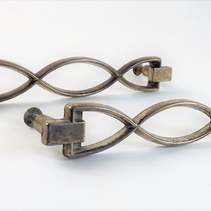 May include: Two antique-style cabinet pulls with a decorative, intertwined design. Each pull is made of a tarnished, metallic material and features a rectangular base and a rounded knob. The pulls are designed to be mounted on a cabinet or drawer.