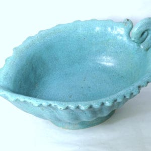 May include: A turquoise ceramic leaf-shaped bowl with a textured surface. The bowl has a scalloped edge and a decorative curled handle. The bowl is elevated on a small, rounded base.