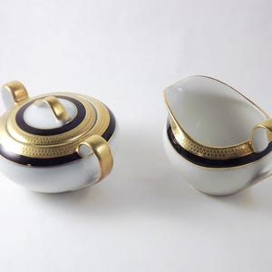 May include: A white porcelain sugar bowl with a lid and a matching creamer pitcher. Both pieces feature a deep navy blue band, a gold rim, and a gold geometric Greek key pattern. The sugar bowl has two gold handles and a gold lid handle.