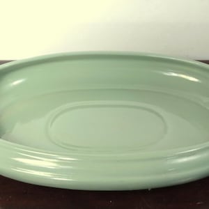 May include: A light green, oval-shaped ceramic serving dish. The dish has a wide, rounded rim and a shallow interior. The color is a soft, muted green, and the dish appears to be in good condition. It is likely used for serving food or as a decorative piece.