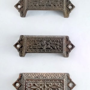 Pode incluir: Três puxadores de gaveta antigos de bronze com desenhos florais e geométricos ornamentados. Cada puxador tem um corpo retangular com extremidades decorativas e dois orifícios de montagem. Os puxadores estão dispostos verticalmente sobre um fundo branco.