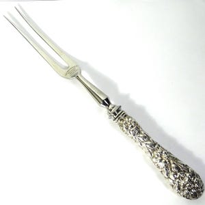 May include: A silver carving fork with two tines. The handle is intricately detailed with floral patterns. The fork has a shiny, reflective surface, and the handle is slightly curved. The image is taken on a white background.