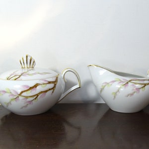 May include: A white porcelain sugar bowl with a lid and a matching creamer pitcher. Both pieces feature a hand-painted design of brown branches with soft pink and green buds, accented by thin gold trim on the handles, rims, and lid finial.