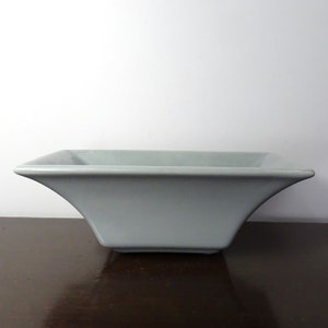 Op de afbeelding: Een lichtgrijze, vierkante keramische kom. De kom heeft een uitlopend, gebogen ontwerp aan de zijkanten en staat op een donkerbruin oppervlak. De kom is leeg en lijkt nieuw.