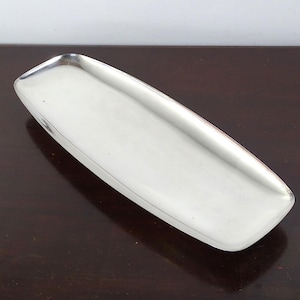 May include: A rectangular silver-colored metal tray with rounded corners and slightly raised edges. The surface of the tray is smooth and reflective, featuring a polished finish that catches the light across its elongated, shallow basin design.