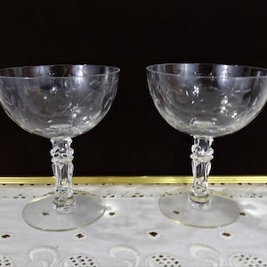 May include: Two clear champagne coupe glasses with etched floral designs. The glasses have a rounded bowl, a decorative stem, and a circular base. The background is dark, and the glasses are on a white surface.