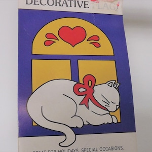 May include: Decorative flag featuring a white cat with a red bow, sleeping in front of a yellow window with a red heart and accents on a blue background. The flag is designed for holidays and special occasions.