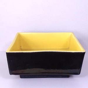 May include: A rectangular ceramic planter with a black exterior and a bright yellow interior. The planter has a square base and a slightly flared rim. The contrast of colours creates a modern, eye-catching design.