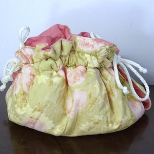 May include: A floral drawstring bag with a yellow and pink rose pattern. The bag has a pink interior and white drawstring cords. The fabric is gathered at the top, creating a ruffled effect. The bag is designed to hold small items.