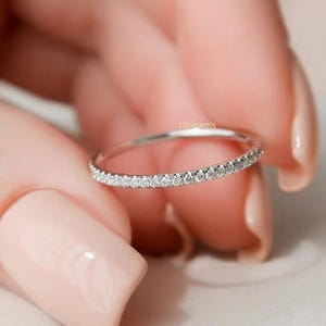Puede incluir: Un delicado anillo de plata con una banda de pequeñas piedras claras y brillantes. El anillo se sostiene entre los dedos con esmalte de uñas rosa claro.