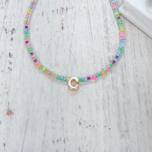 May include: A necklace featuring a strand of small, multicolored glass beads in shades of pink, blue, green, orange, and purple. A single, gold-toned circular charm with the letter C embossed in the center hangs from the bottom of the strand.