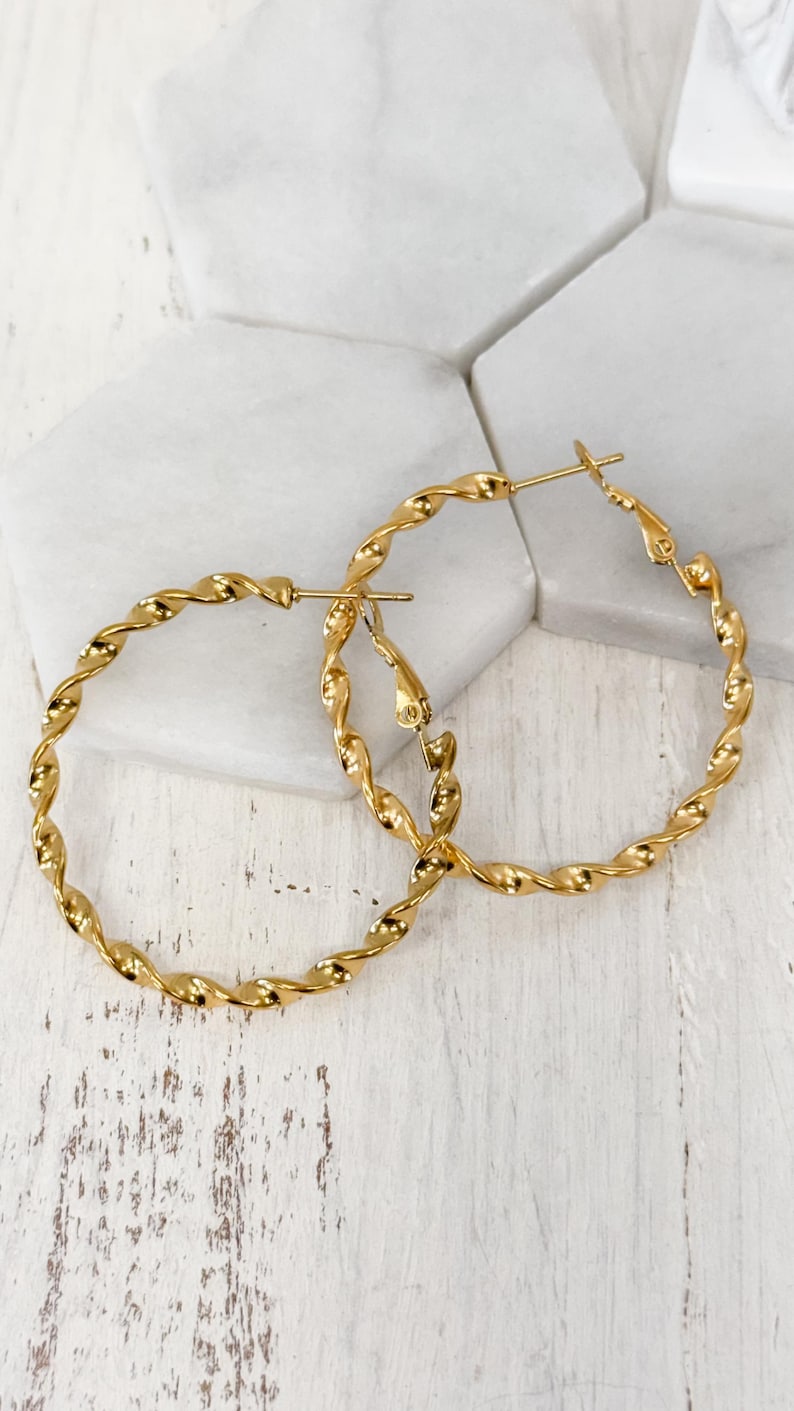 May include: A pair of gold-toned hoop earrings featuring a twisted, rope-like texture. The earrings are circular in shape and include a hinged post closure mechanism at the top, designed to secure the hoop when worn through the earlobe.