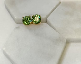 May include: A pair of gold-colored stud earrings featuring round, light green gemstones. The earrings are set on a white, hexagonal surface. The gemstones have a faceted appearance, reflecting light.