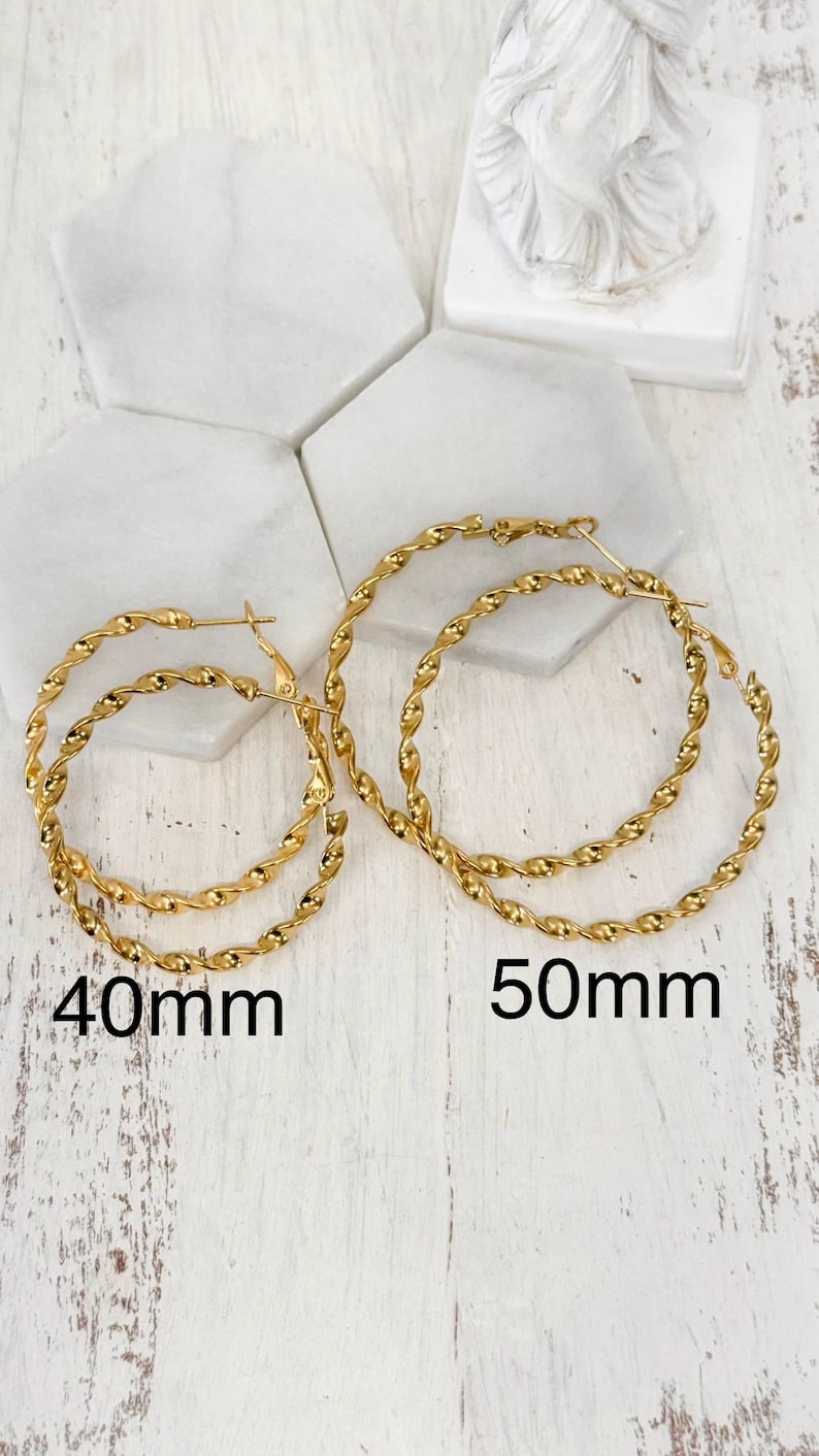 May include: Two pairs of gold-toned hoop earrings with a twisted rope texture. One pair is labelled 40mm and the other pair is labelled 50mm. The earrings feature a circular design with a hinged post closure, showcasing a polished metallic finish.