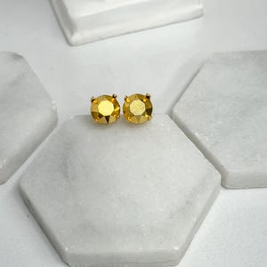 May include: A pair of gold-toned stud earrings with large, faceted, yellow-gold colored stones. The earrings are displayed on a white marble surface, with a small white statue in the background. The earrings are a jewelry item.