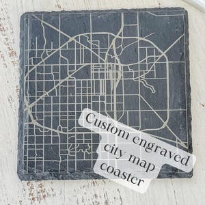 May include: A square, dark gray slate coaster with a detailed, engraved city map design. The text "Custom engraved city map coaster" is partially visible. The coaster is on a white wooden surface.