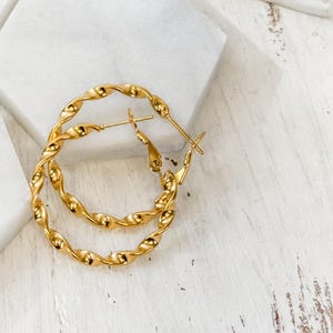 May include: A pair of gold-toned hoop earrings featuring a twisted, rope-like texture. The earrings are circular in shape and include a standard post-back closure mechanism, showcasing a polished, reflective metallic finish throughout the design.