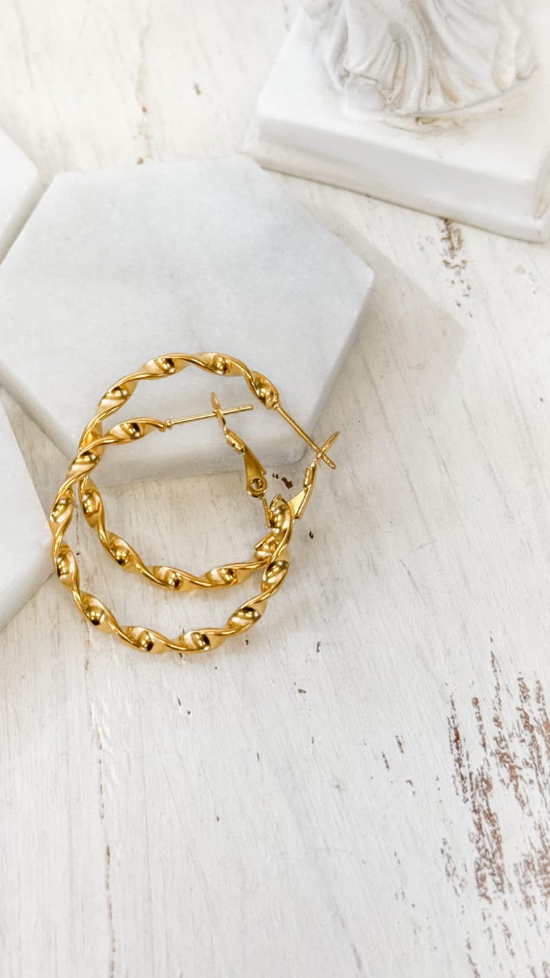 May include: A pair of gold-toned hoop earrings featuring a twisted, rope-like texture. The earrings are circular in shape and include a standard post-back closure mechanism, showcasing a polished, reflective metallic finish throughout the design.