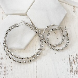 May include: Two pairs of silver hoop earrings with a twisted rope design. One pair is larger than the other. The earrings are displayed on a white wooden surface with white hexagonal tiles in the background.