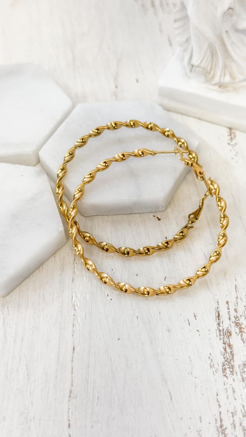 May include: A pair of gold-toned hoop earrings featuring a continuous twisted rope texture. The circular earrings are constructed from a polished metal wire that forms a spiral pattern throughout the entire circumference of each hoop.