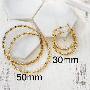 May include: Two pairs of gold-toned hoop earrings with a twisted rope texture. One pair is labelled 50mm and the other pair is labelled 30mm. The earrings feature a thin wire post closure and are arranged on a white surface with hexagonal tiles.