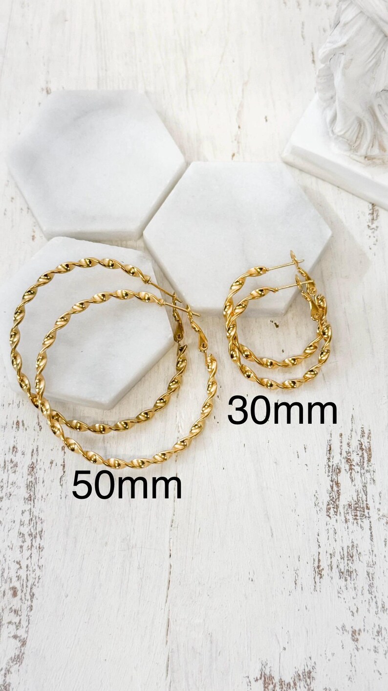 May include: Two pairs of gold-toned hoop earrings with a twisted rope texture. One pair is labelled 50mm and the other pair is labelled 30mm. The earrings feature a thin wire post closure and are arranged on a white surface with hexagonal tiles.