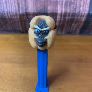 May include: A monkey-faced PEZ dispenser with a tan head, black facial features, and blue eyes. The dispenser has a bright blue stem and base. The background is a wooden surface.