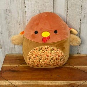 May include: A plush turkey toy with a brown body, orange head, and a patterned belly. The turkey has black eyes, a yellow beak, and a red wattle. The toy is designed to look like a cartoon turkey.