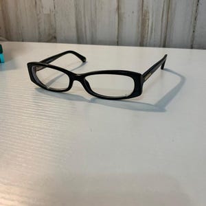 May include: Black rectangular eyeglasses with clear lenses and thin frames. The glasses feature a glossy finish on the plastic rims and temples, with a simple bridge connecting the two lenses. The design is sleek and functional for daily vision needs.