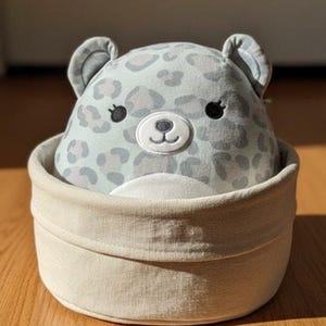 May include: A plush toy shaped like a leopard head with light blue and grey spots, black embroidered eyes, and a white muzzle, nestled inside a round, beige fabric storage basket with a folded rim and a simple, clean design.
