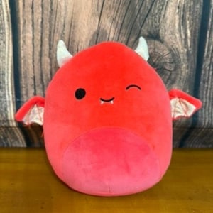 May include: A red devil-shaped plush toy with small white horns, a winking eye, and small wings. The toy has a lighter red belly and a smiling mouth. The background is a wooden surface.