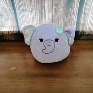 以下が含まれることがあります： 大きな耳と小さな花が付いた、ラベンダー色の象の頭のぬいぐるみ。象は黒い目とステッチされた鼻を持っています。おもちゃは木製の表面にあります。