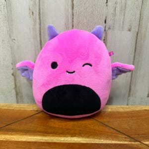 May include: A pink plush toy shaped like a winking creature with purple horns and wings. It has a black belly and eyes. The background is a wooden surface.
