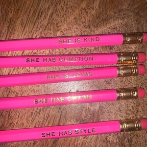 Puede incluir: Cinco lápices de color rosa brillante con letras y férulas doradas. Cada lápiz tiene una afirmación positiva diferente: "SHE IS KIND", "SHE HAS GUMPTION", "SHE IS LOYAL", "SHE HAS CHARM" y "SHE HAS STYLE".