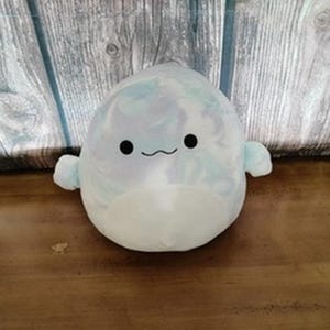 May include: A plush, round, light blue stuffed animal with small arms and a smiling face. The toy has a white belly and a marbled pattern on its head and back. The background is a wooden surface.